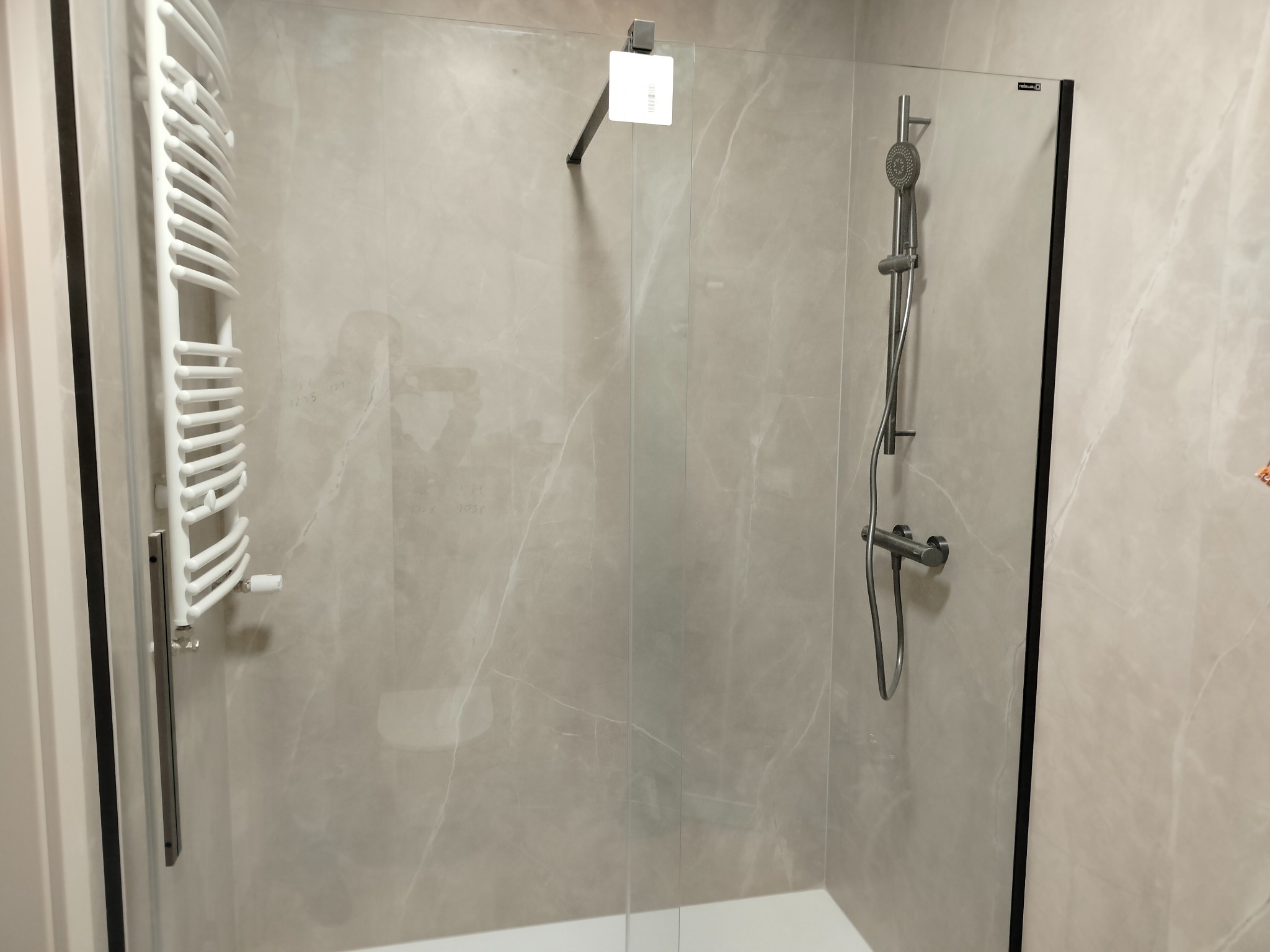 Kabina prysznicowa z szarą marmurową okładziną, wyposażona w biały grzejnik drabinkowy, deszczownicę i słuchawkę prysznicową.