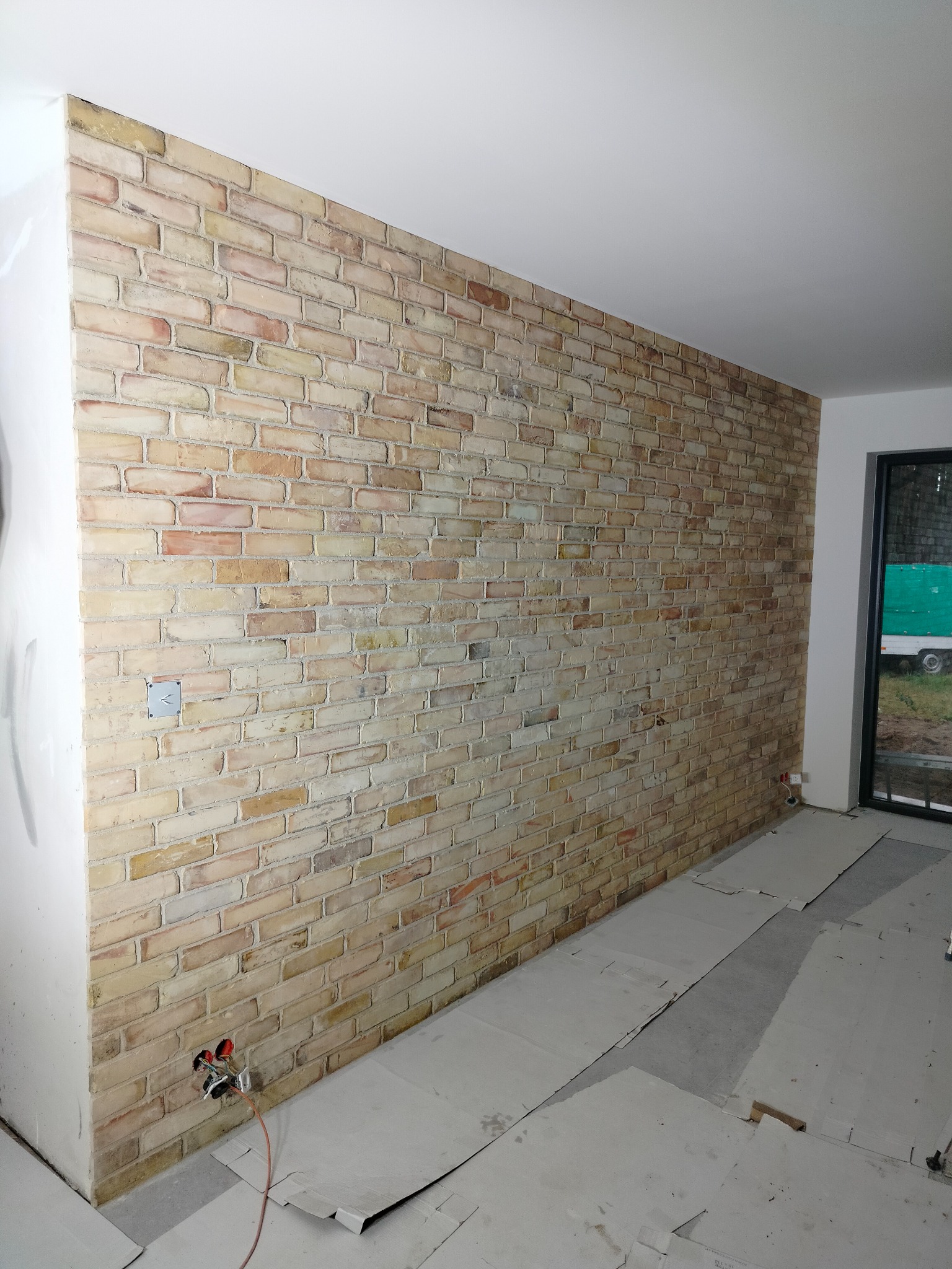 Ściana wykończona cegłą dekoracyjną w odcieniach beżu i czerwieni, widoczne przygotowania do instalacji elektrycznej, podłoga zabezpieczona kartonem.