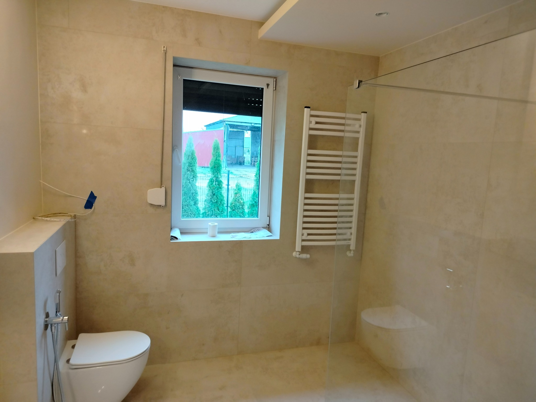 Wykończona łazienka w Pile z beżowymi płytkami imitującymi kamień, widoczny biały sedes z bidetką, grzejnik drabinkowy i przeszklona kabina prysznicowa.