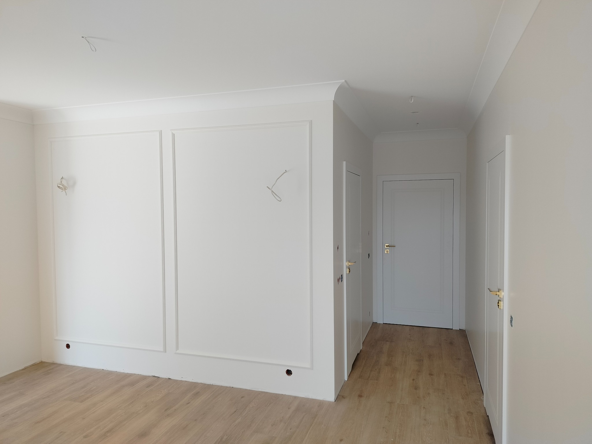 Białe, nowocześnie wykończone wnętrze z panelami podłogowymi, ozdobnymi listwami ściennymi i dwiema białymi drzwiami z złotymi klamkami, z widocznymi otworami na gniazdka elektryczne i przewodami...