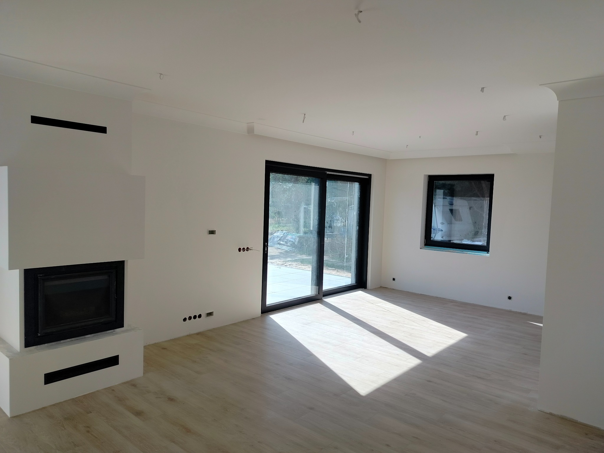 Jasne, przestronne wnętrze z kominkiem, dużymi przesuwnymi drzwiami balkonowymi i panelami podłogowymi, widoczne niedokończone punkty elektryczne na ścianach.
