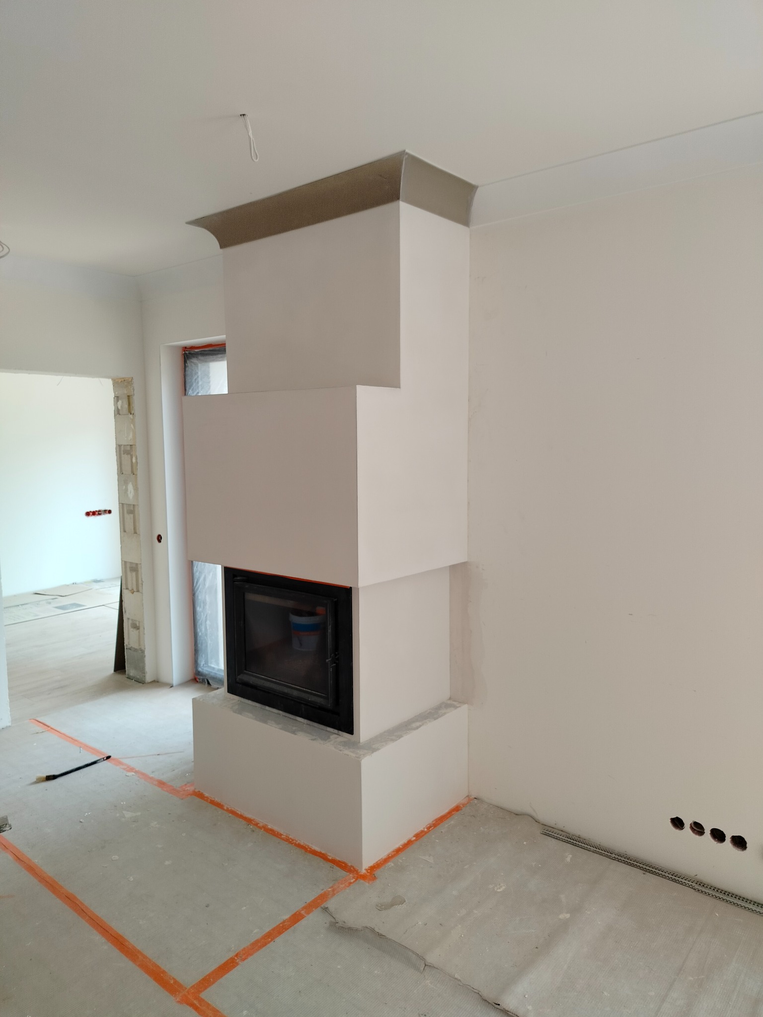 Wnętrze w trakcie prac wykończeniowych: obudowa kominka z prostokątnymi elementami, listwy dekoracyjne pod sufitem, pomarańczowe linie wyznaczające obszar prac na podłodze, widoczne przewody...