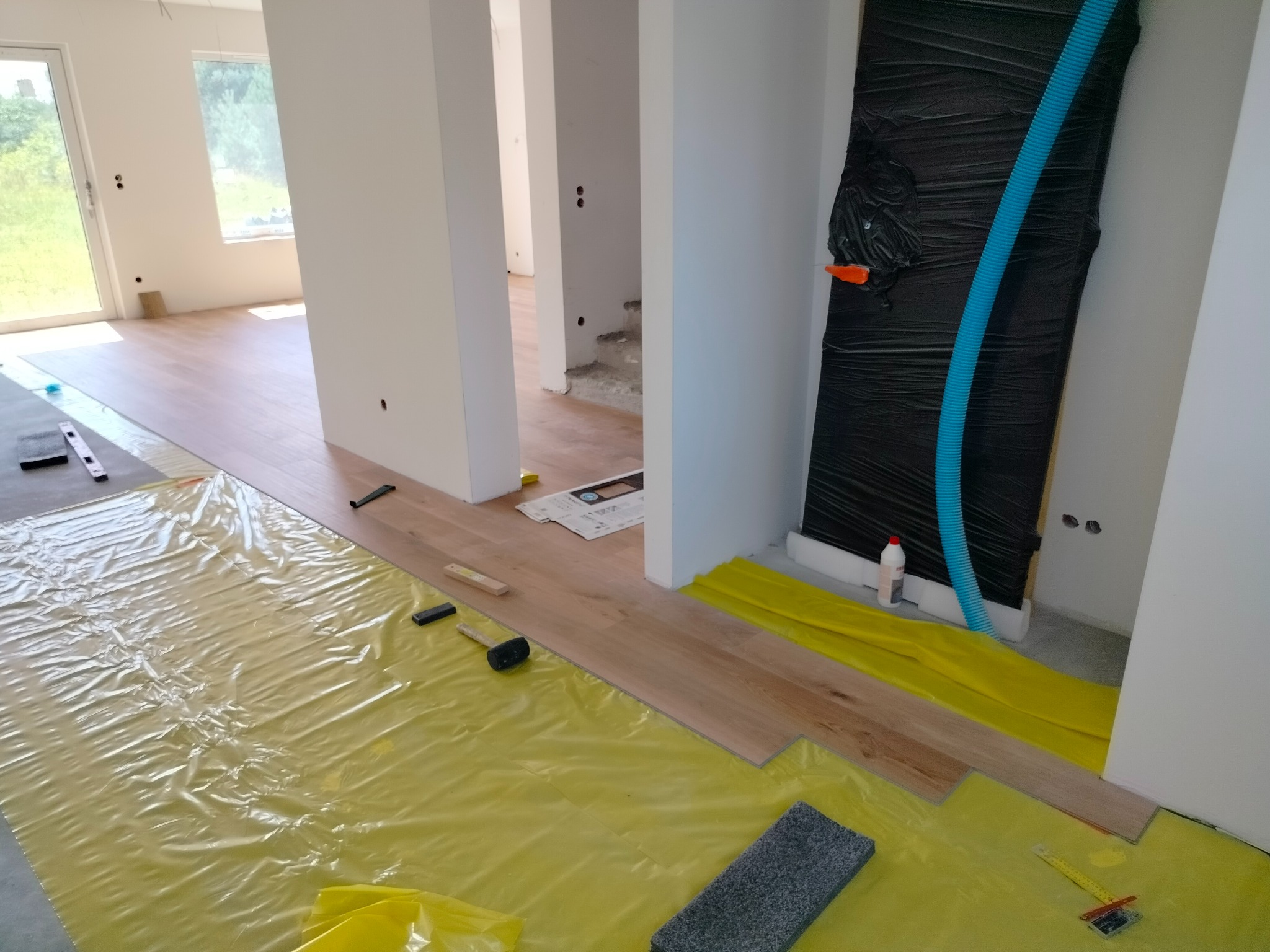 Montaż paneli podłogowych w trakcie realizacji, widoczna folia ochronna, gumowy młotek, listwy i przycięte panele, ściany przygotowane do dalszych prac wykończeniowych.