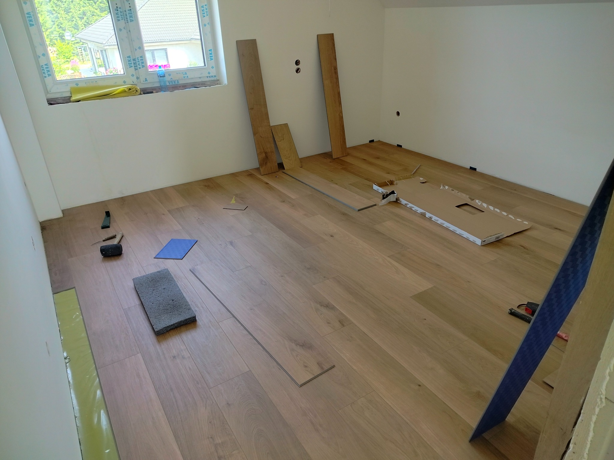 Układanie jasnych paneli podłogowych w pokoju, widoczne narzędzia, fragment okna z widokiem na dom, płyta dociskowa i kilka nieułożonych desek.