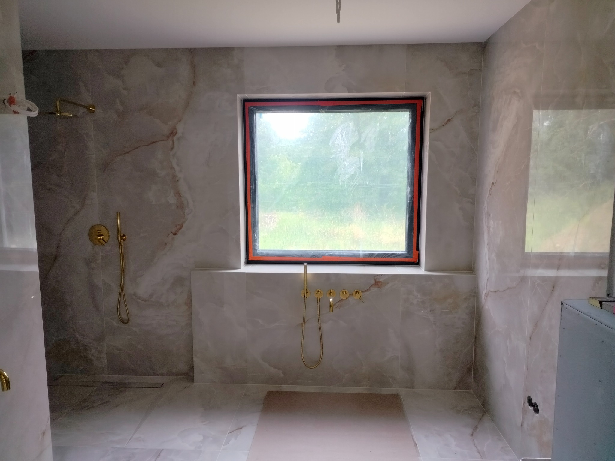 Wykończona łazienka z marmurowymi płytkami, złotą armaturą i oknem z widokiem na zieleń. Uwagę zwraca minimalistyczny design i naturalne światło wpadające przez okno.