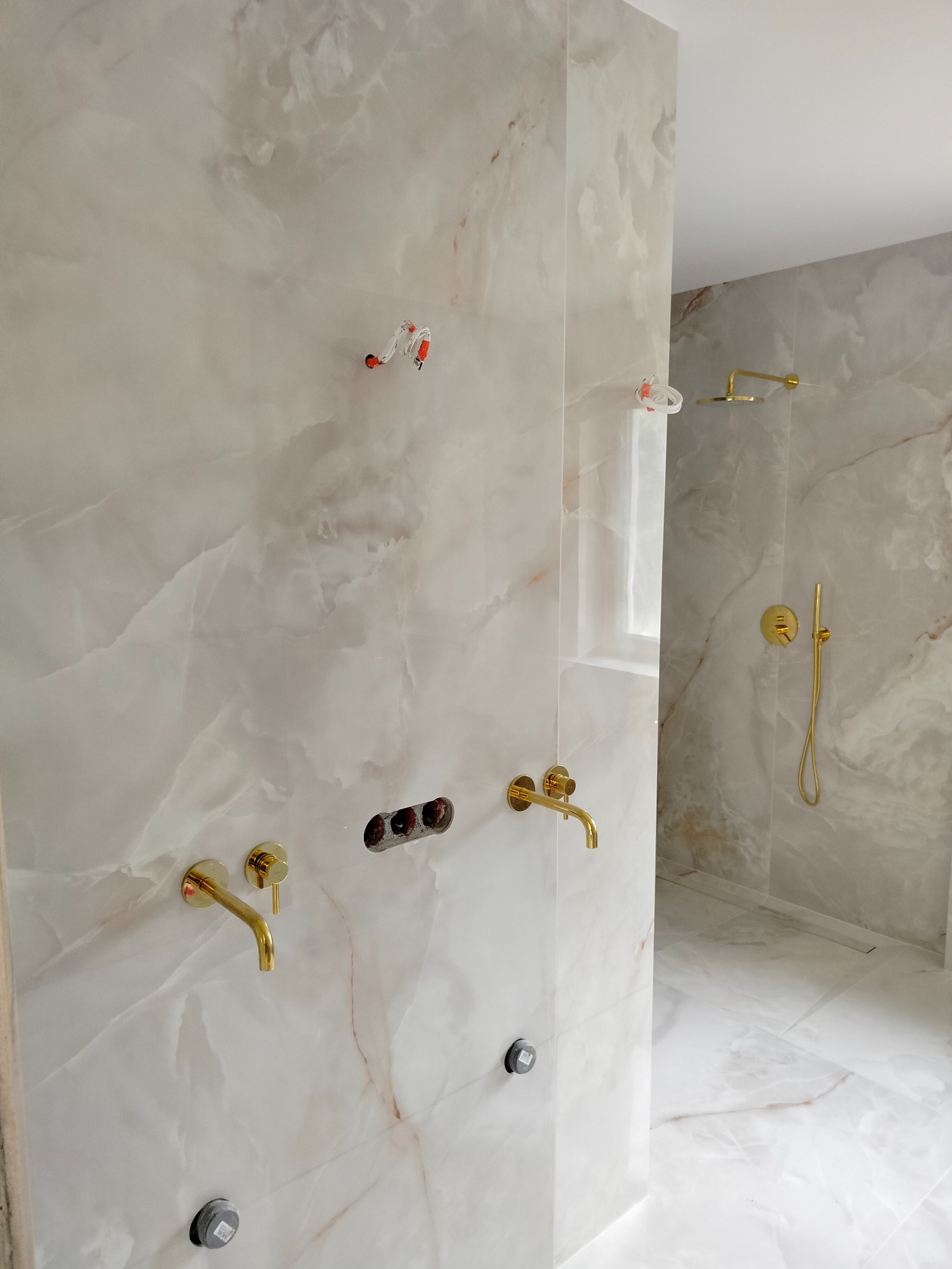 Montaż złotej armatury łazienkowej na ścianie z płytek imitujących marmur, widoczne przygotowane punkty instalacyjne