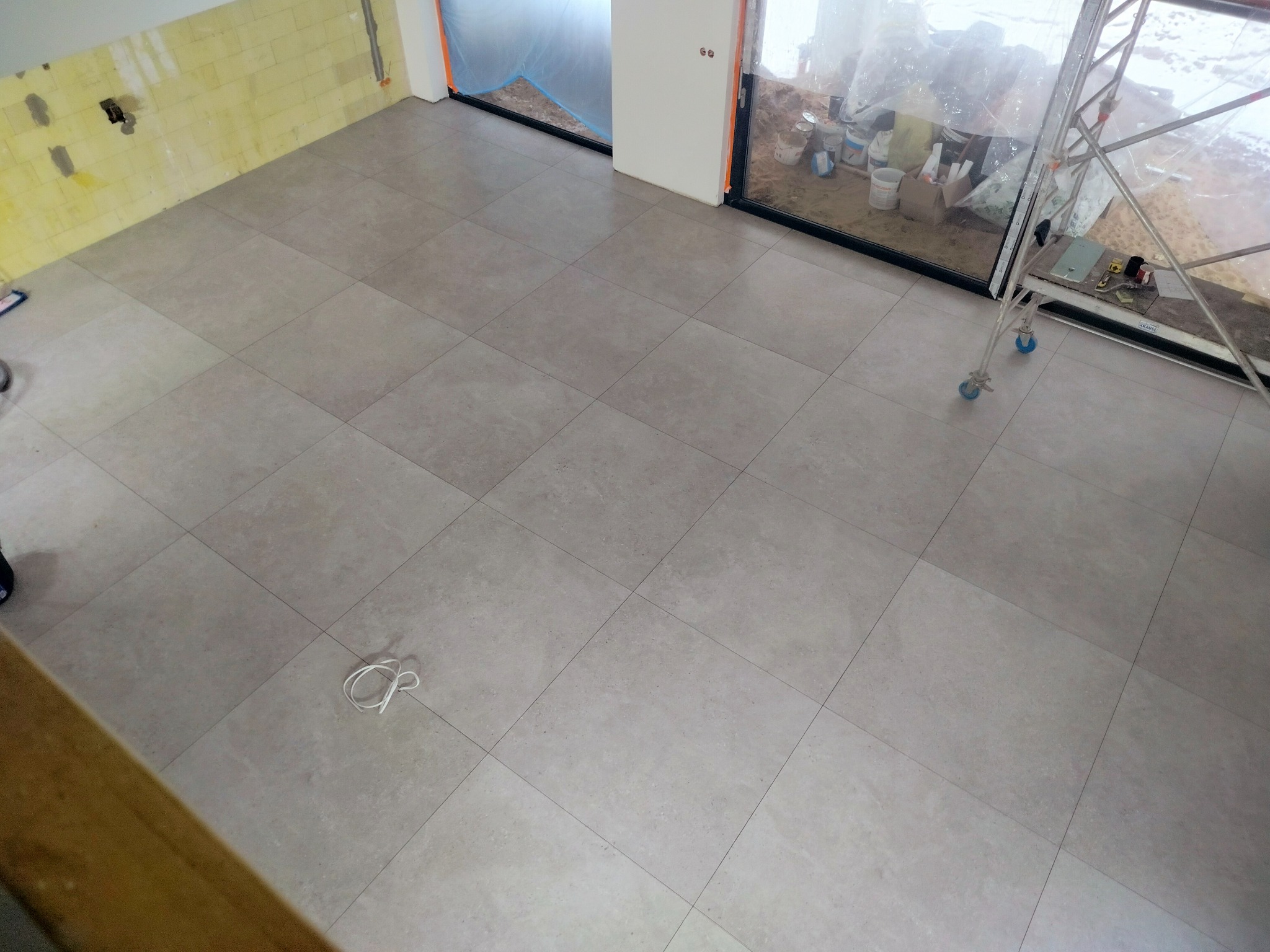 Ułożone szare płytki podłogowe w nowym pomieszczeniu, widoczny fragment żółtej ściany w trakcie prac wykończeniowych i rusztowanie przy oknie.