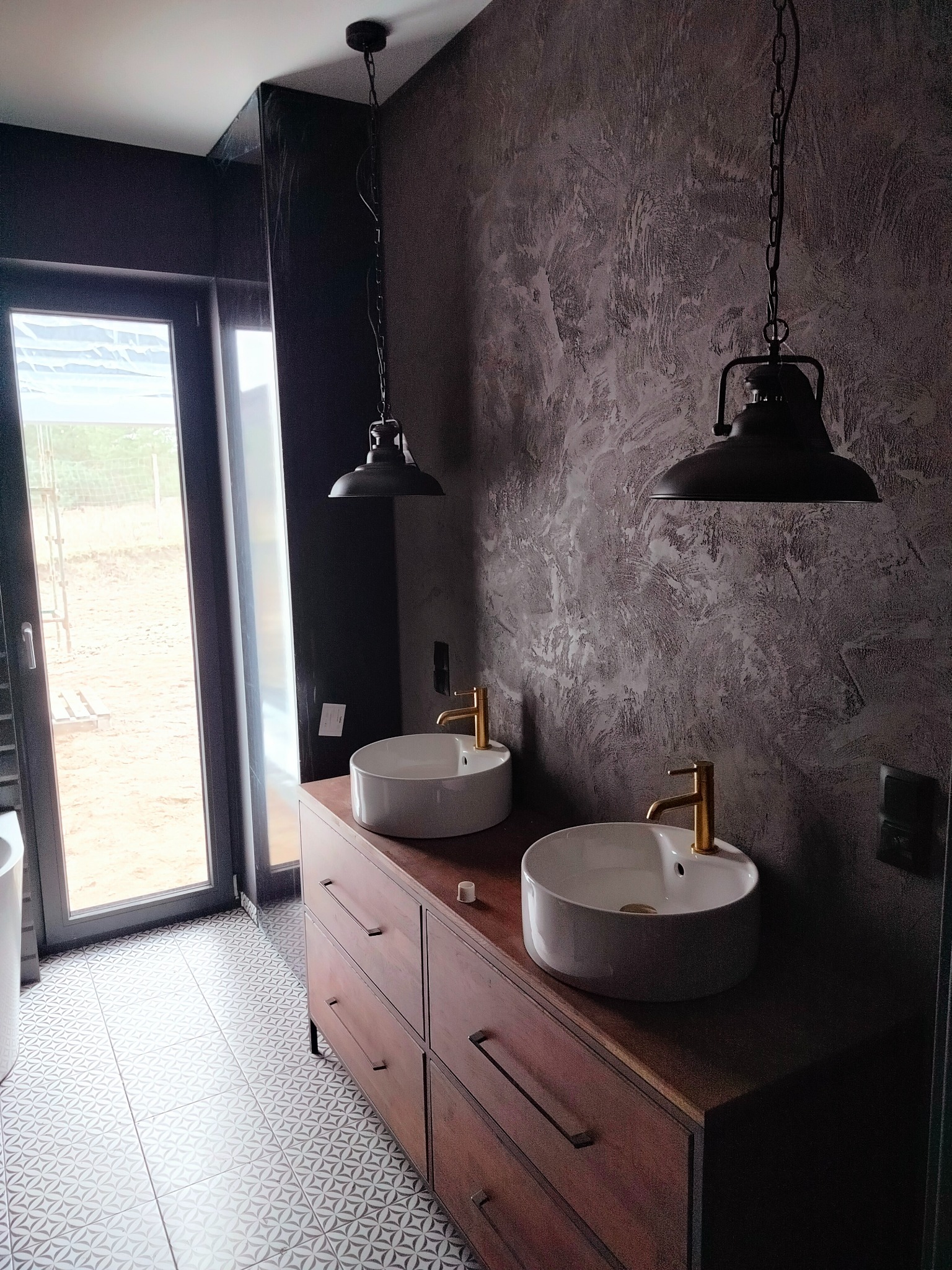 Nowoczesna łazienka z dwoma okrągłymi umywalkami nablatowymi z złotymi bateriami, drewnianą szafką z szufladami, dekoracyjną ścianą z efektem betonu oraz wiszącymi lampami w stylu industrialnym.