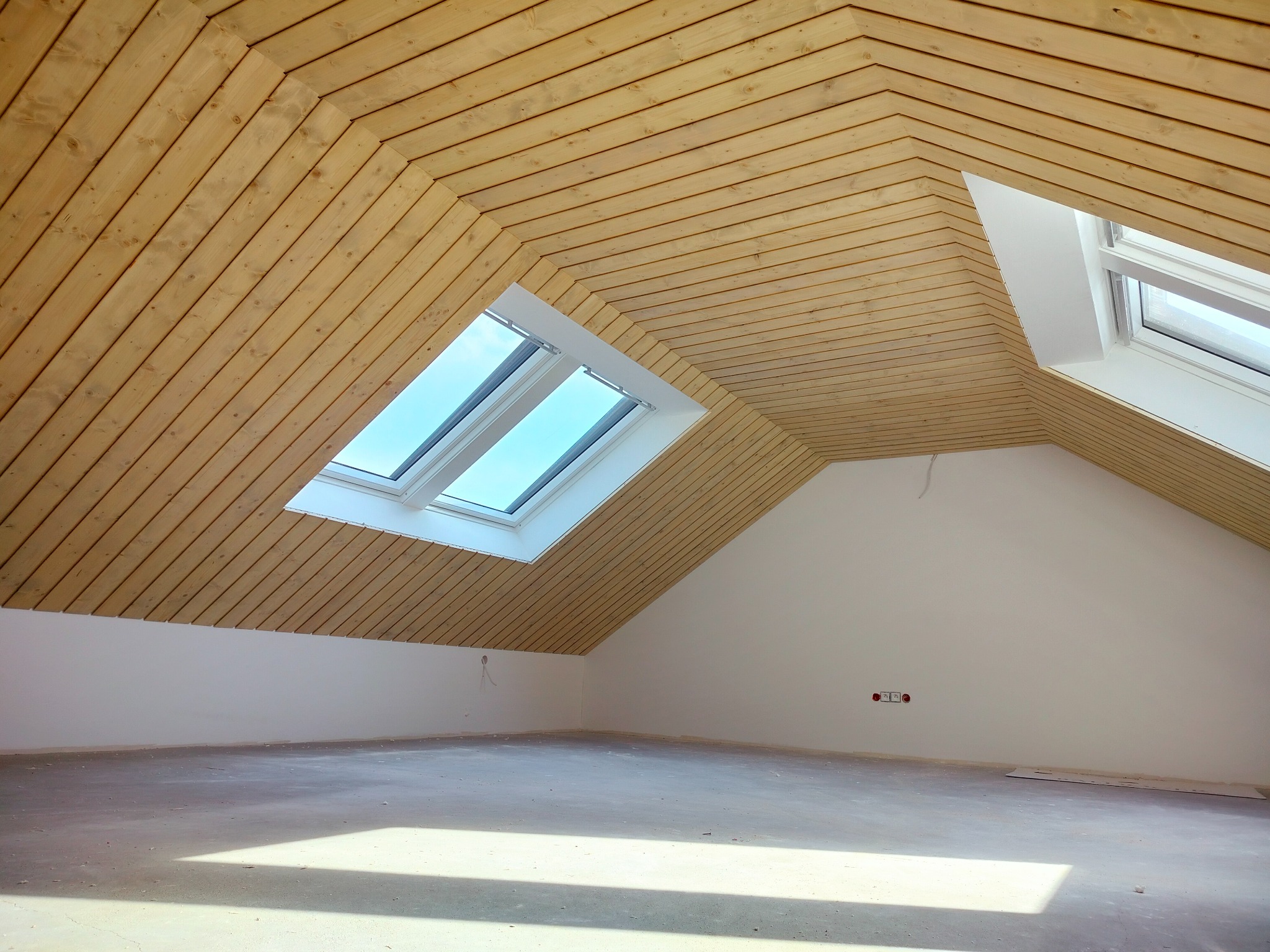 Poddasze z drewnianą boazerią na skośnym suficie i dwoma oknami dachowymi, ściany pomalowane na biało, surowa betonowa podłoga, widoczne gniazdka elektryczne.