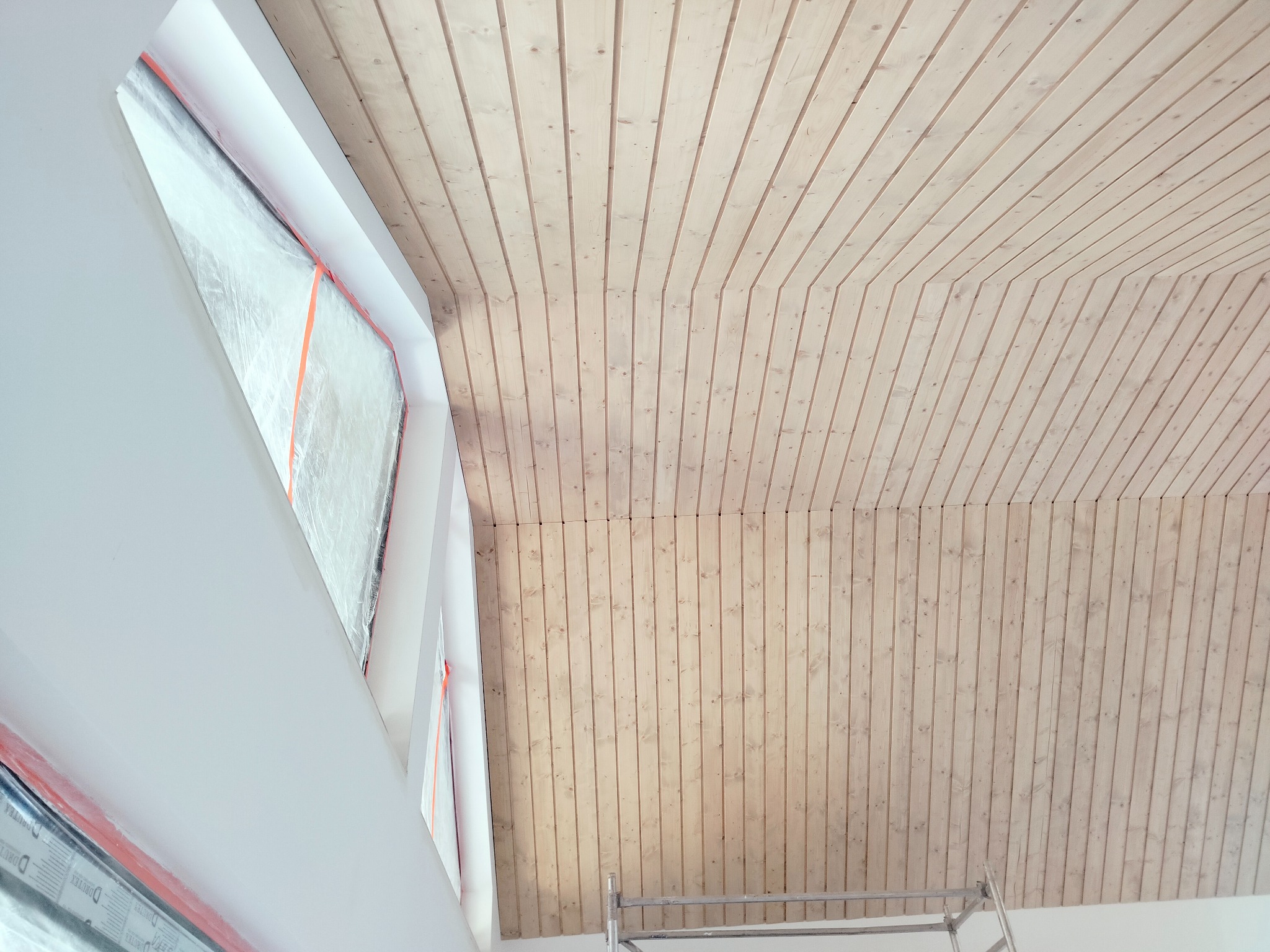 Drewniana boazeria na skośnym suficie z oknem dachowym w trakcie montażu, zabezpieczonym folią. Fragment drabiny malarskiej widoczny w rogu.
