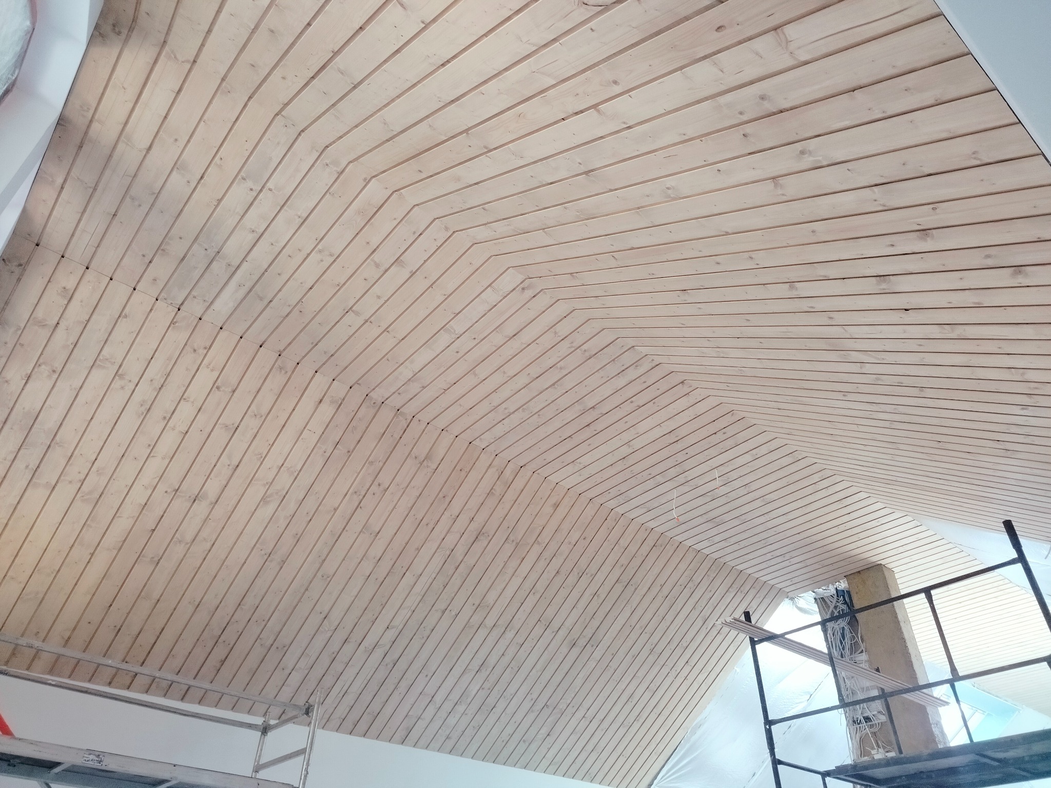 Wykończony sufit poddasza z drewnianą boazerią w jasnym odcieniu, widoczne rusztowanie i folia ochronna, skierowane pod kątem ujęcie podkreśla geometrię ułożenia desek.