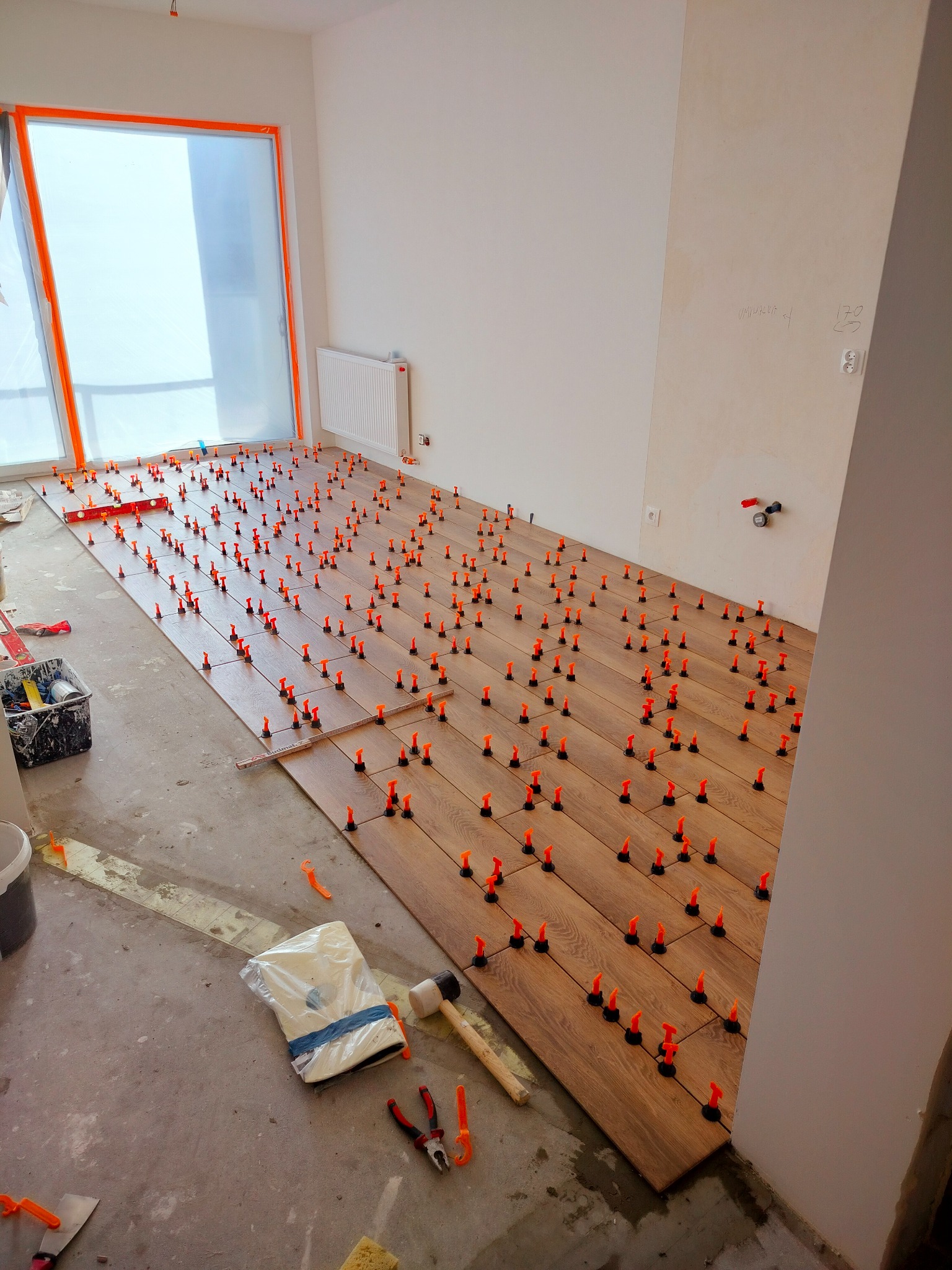 Układanie drewnopodobnych paneli podłogowych z użyciem pomarańczowych klipsów poziomujących, widoczne narzędzia i materiały na podłodze, okno zabezpieczone folią.