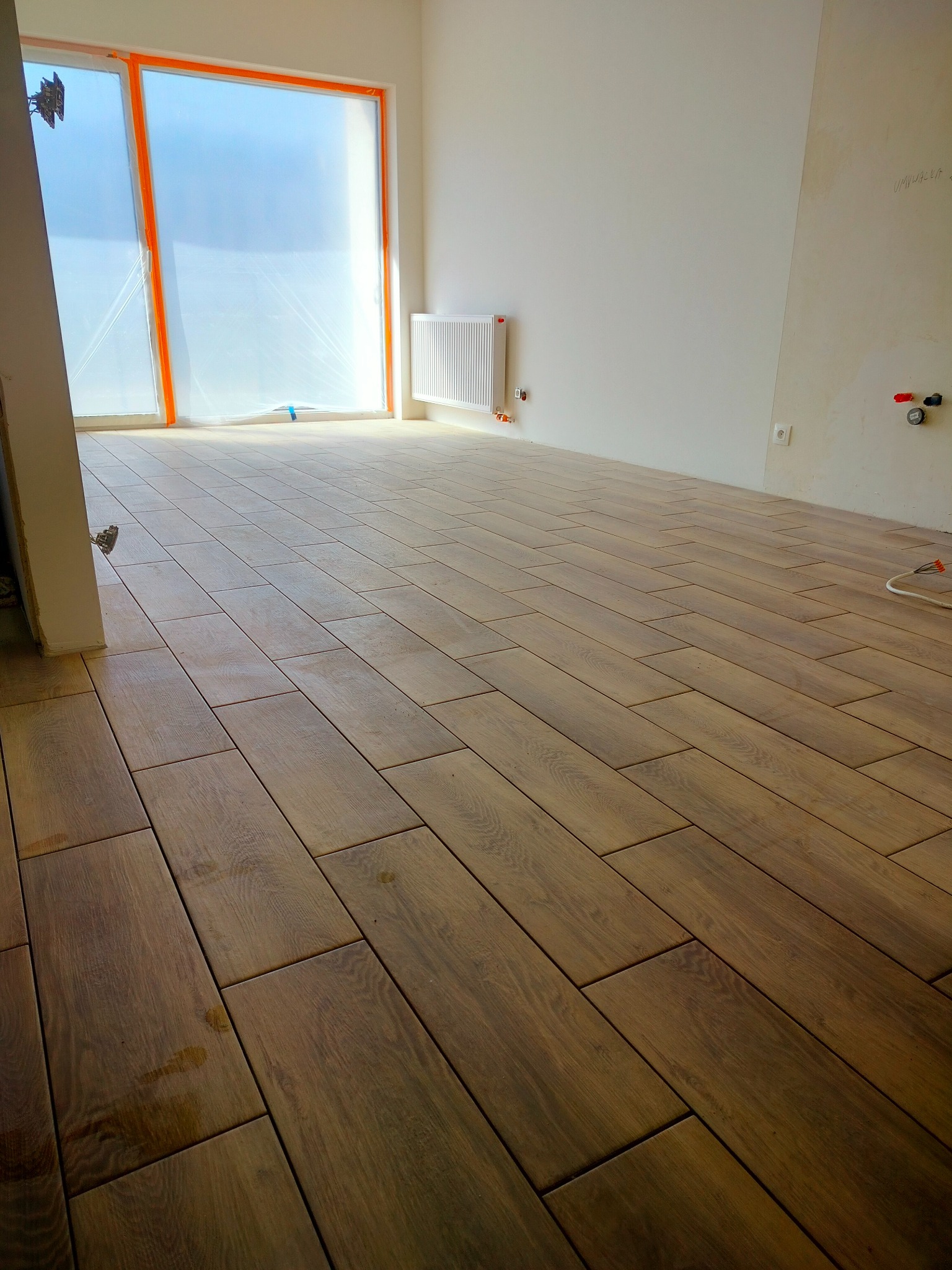 Wnętrze w trakcie prac wykończeniowych: ułożona podłoga z paneli imitujących drewno, okno zabezpieczone folią malarską, grzejnik na ścianie i widoczne instalacje elektryczne.