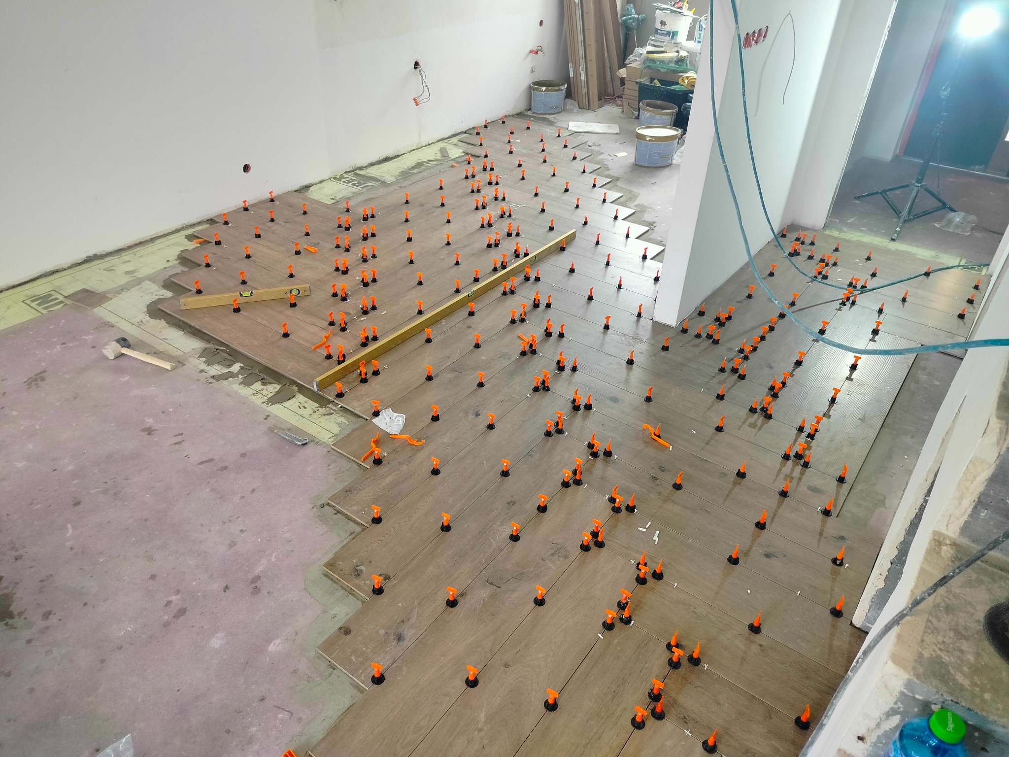 Montaż paneli podłogowych w jodełkę, z użyciem systemu poziomowania z pomarańczowymi klipsami, widoczna poziomica i fragment surowej podłogi.