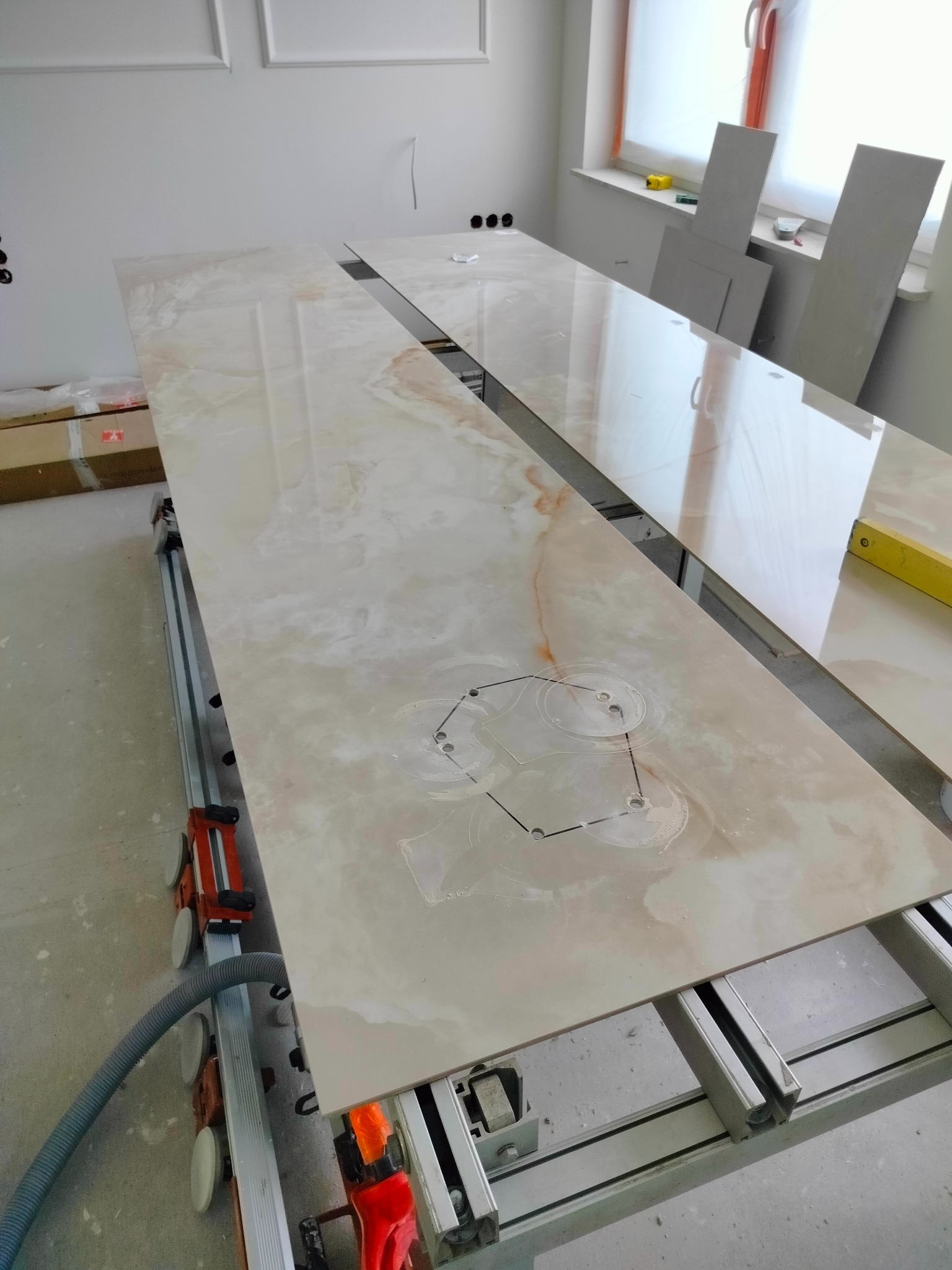 Przygotowanie blatu z płyty imitującej marmur z wyciętymi otworami na zlew, na stole do cięcia płytek, w tle okno i fragment ściany z dekoracyjną sztukaterią.