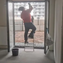 Mężczyzna na drabinie czyści okno balkonowe w surowym wnętrzu, widok na teren zewnętrzny z gruzem i nowym budownictwem w tle.