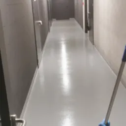 Mop z niebieskimi rękawicami czyści błyszczącą, szarą podłogę w długim korytarzu z szarymi ścianami z bloków, rurami pod sufitem i ciemnymi drzwiami.