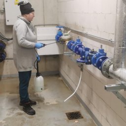 Martyna Klimczak Firma Sprzątająca - Kobieta w rękawicach ochronnych spryskuje instalację wodną w pomieszczeniu technicznym, widoczne rury, zawory i białe ściany z betonu komórkowego.