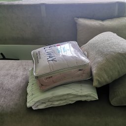 Martyna Klimczak Firma Sprzątająca - Zapakowany komplet pościeli marki ELWAY, w przezroczystym opakowaniu z suwakiem, leży na złożonej, jasnozielonej kołdrze na ciemnoszarej kanapie obok poduszek.