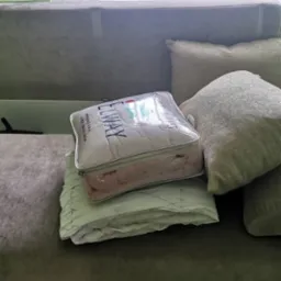 Zapakowany komplet pościeli marki ELWAY, w przezroczystym opakowaniu z suwakiem, leży na złożonej, jasnozielonej kołdrze na ciemnoszarej kanapie obok poduszek.