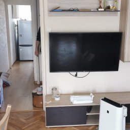 Martyna Klimczak Firma Sprzątająca - Salon w trakcie porządkowania: widoczny telewizor na ścianie, szafka RTV, oczyszczacz powietrza, kartony i torba na podłodze, oraz otwarte drzwi do kuchni z lodówką.