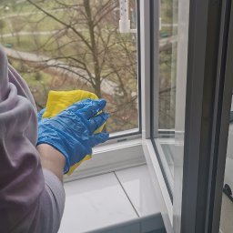 Martyna Klimczak Firma Sprzątająca - Otwarty plastikowy okno z termometrem czyszczone żółtą ścierką przez osobę w niebieskich rękawiczkach, widok na drzewa bez liści za oknem.
