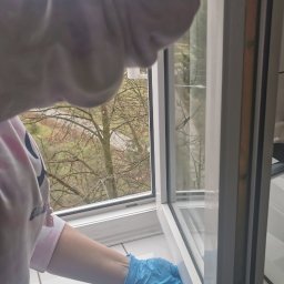 Martyna Klimczak Firma Sprzątająca - Osoba w niebieskich rękawicach czyści ramę okienną od wewnątrz, widok na drzewa za oknem.