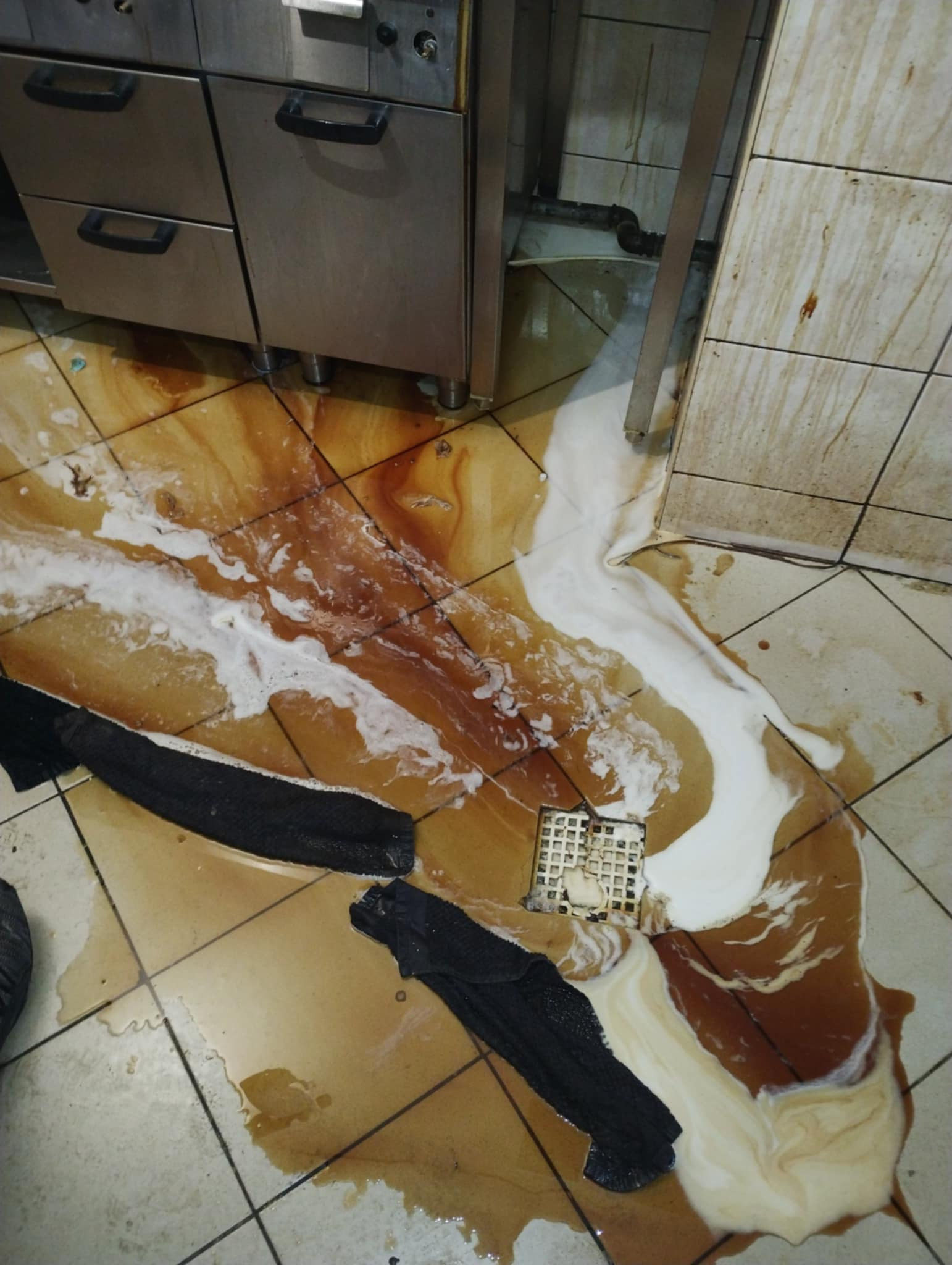 Zalana podłoga w kuchni restauracyjnej w Zduńskiej Woli, widoczne resztki jedzenia i środka czyszczącego, czarne ścierki leżące na brudnej posadzce, odpływ podłogowy częściowo zatkany.