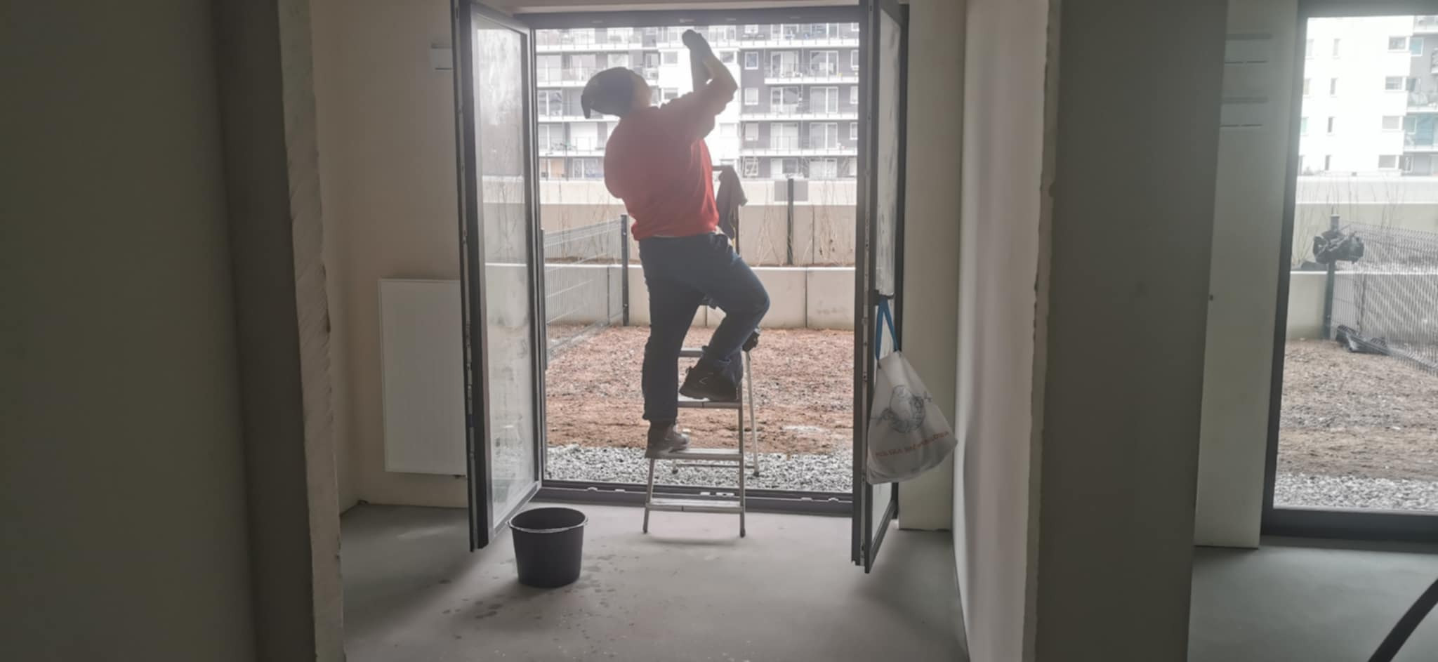 Mężczyzna na drabinie czyści okno balkonowe w surowym wnętrzu, widok na teren zewnętrzny z gruzem i nowym budownictwem w tle.