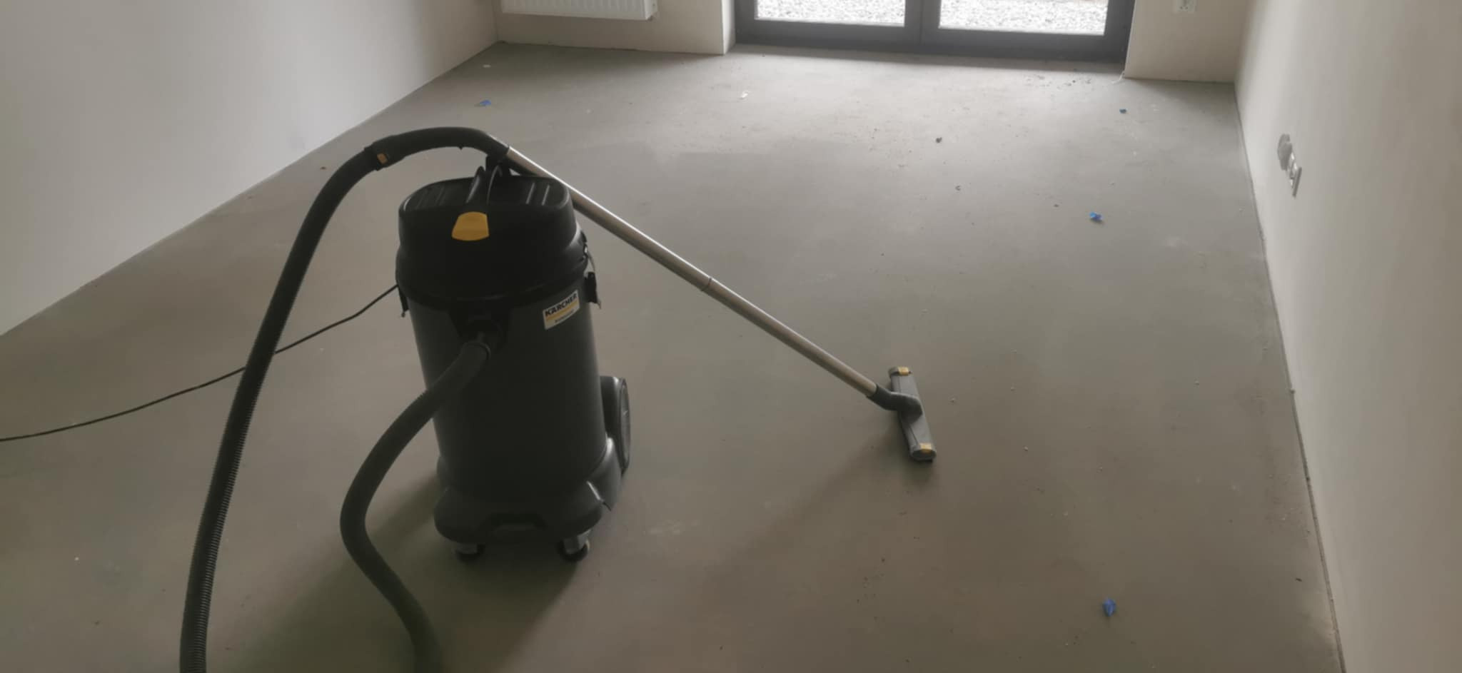 Profesjonalny odkurzacz przemysłowy Karcher w trakcie pracy na szarej, betonowej posadzce w pomieszczeniu po remoncie. Widoczne drobne zabrudzenia i resztki budowlane.