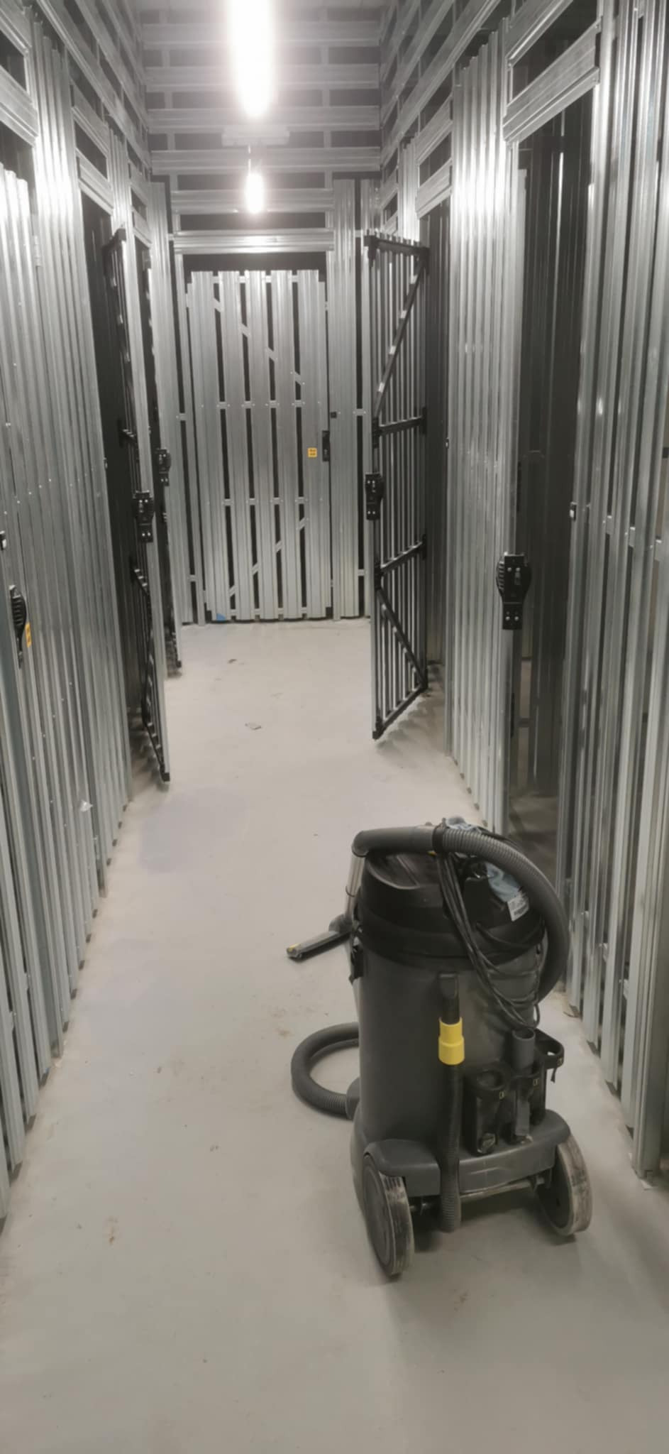 Wnętrze magazynu samoobsługowego w trakcie budowy z widocznym odkurzaczem przemysłowym na posadzce, rzędy metalowych boksów z otwartymi drzwiami.