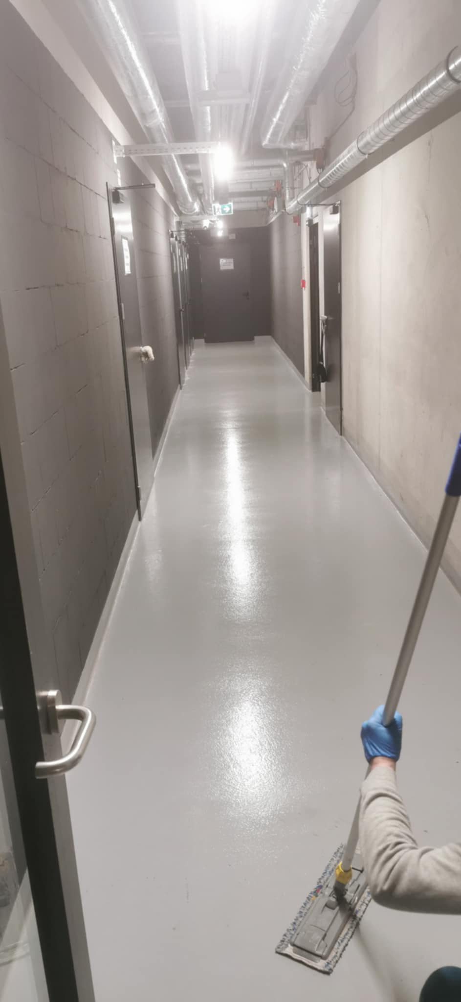 Mop z niebieskimi rękawicami czyści błyszczącą, szarą podłogę w długim korytarzu z szarymi ścianami z bloków, rurami pod sufitem i ciemnymi drzwiami.