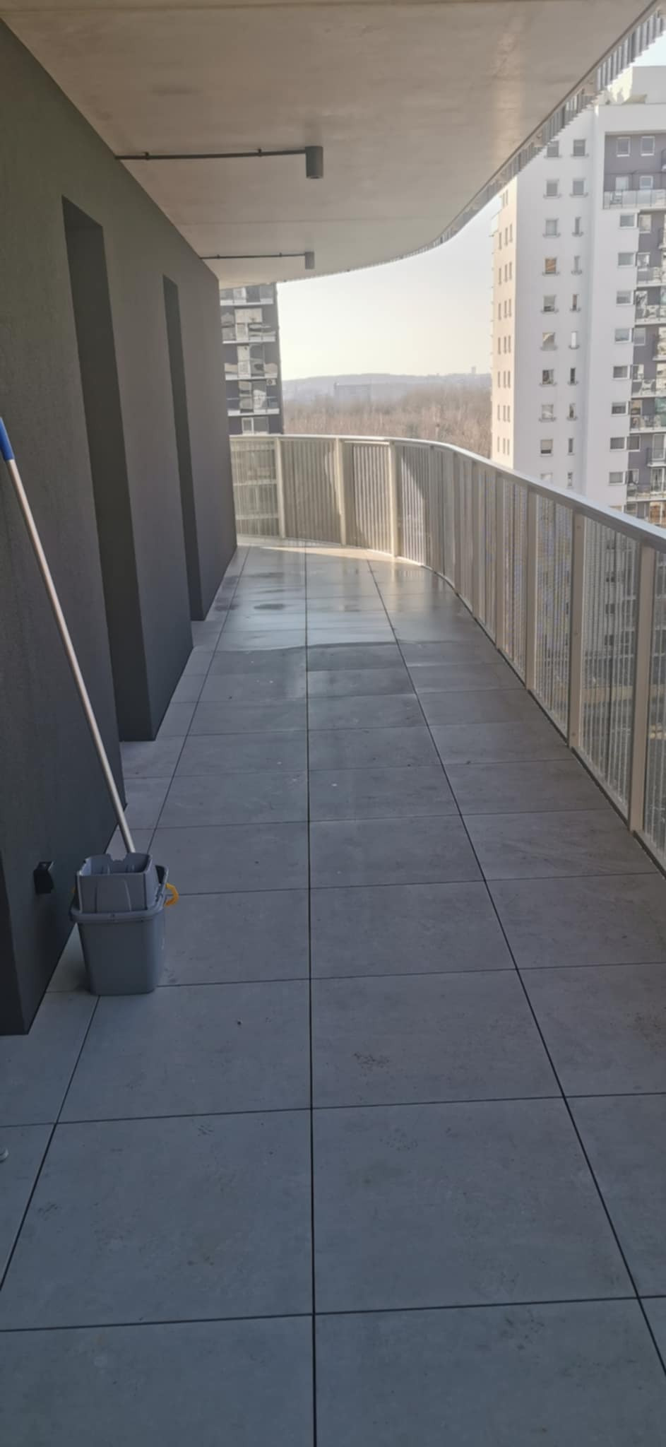 Mop i wiadro na mokrym, wykafelkowanym balkonie apartamentowca z widokiem na miasto w tle.