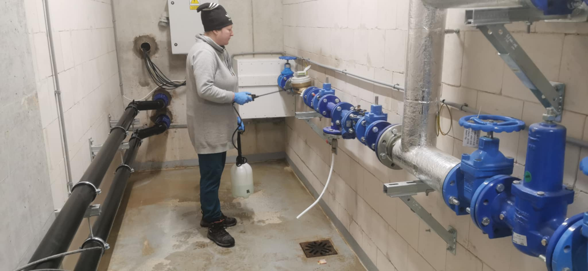 Kobieta w rękawicach ochronnych spryskuje instalację wodną w pomieszczeniu technicznym, widoczne rury, zawory i białe ściany z betonu komórkowego.