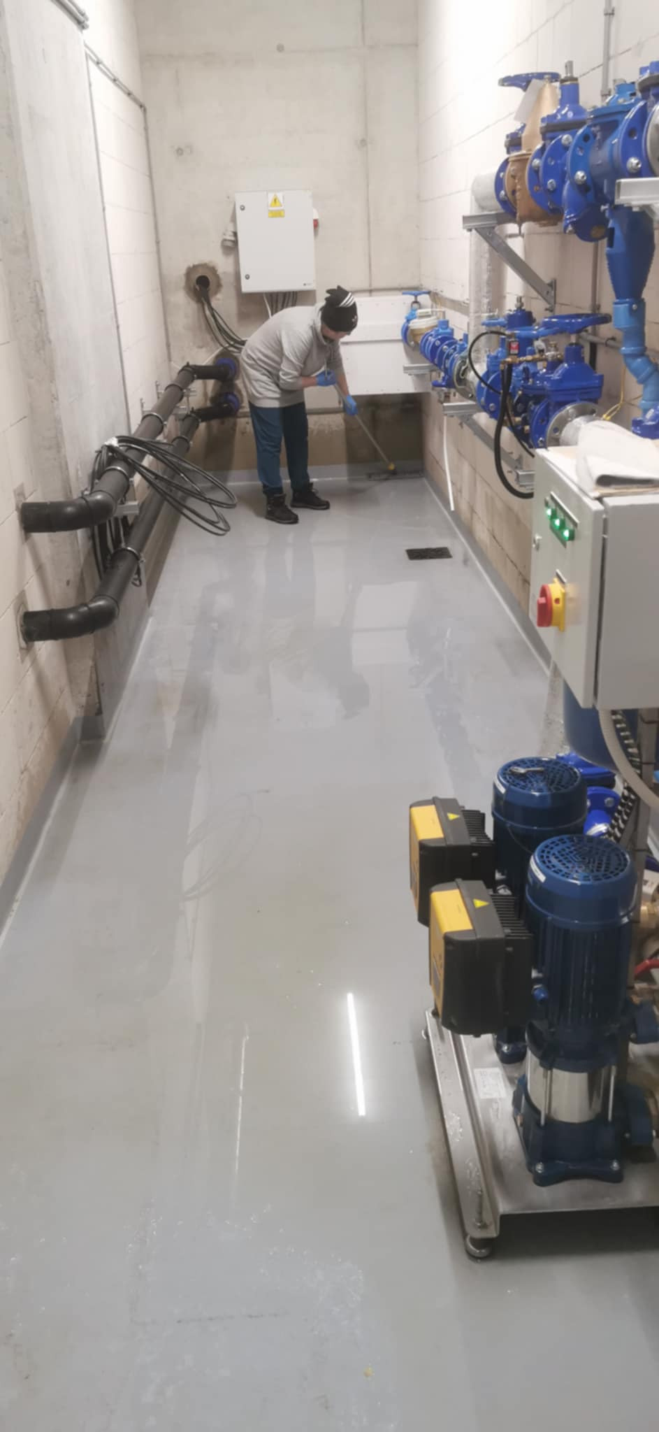 Pracownik w masce i rękawicach myje przemysłową posadzkę w pomieszczeniu z rurami, zaworami i pompami.