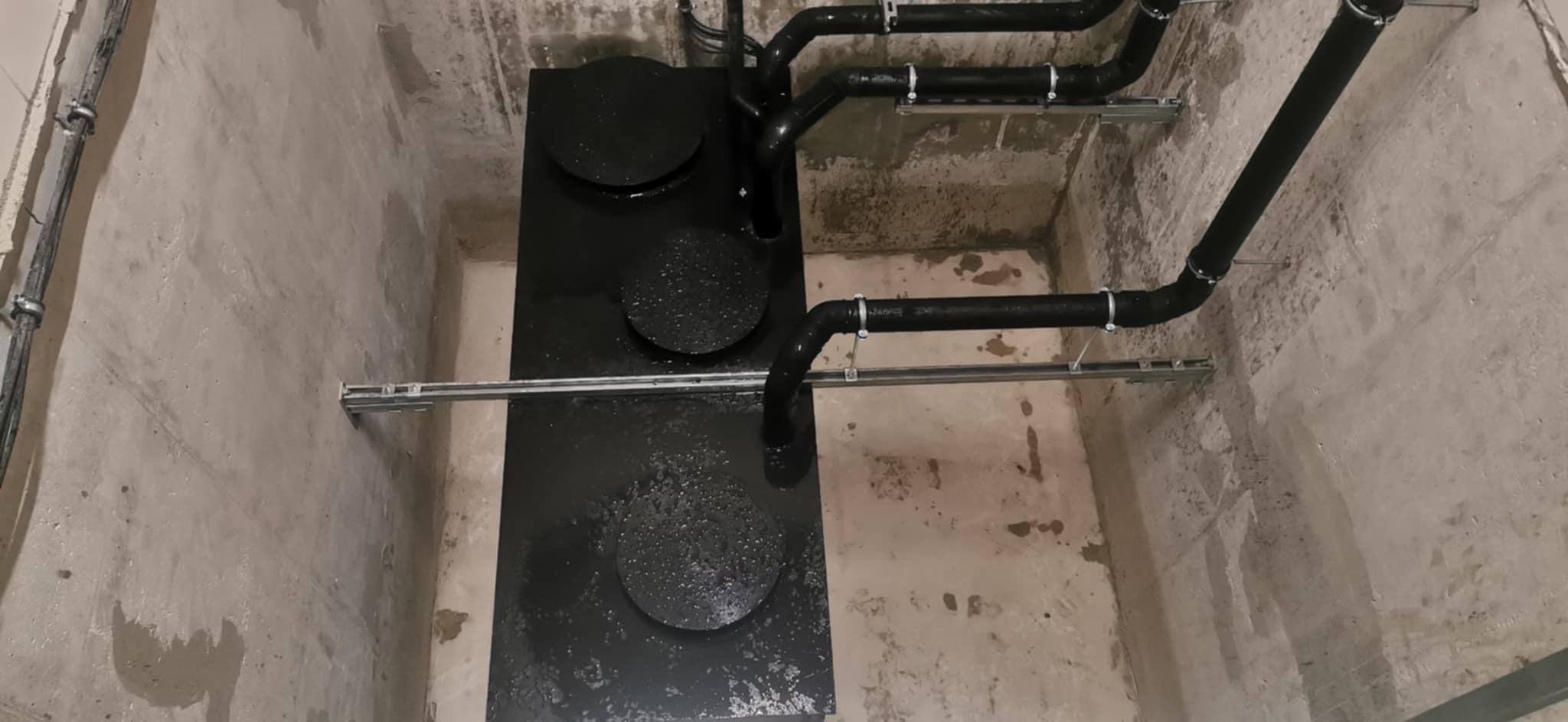 Czarny zbiornik z rurami w betonowym pomieszczeniu, widoczne ślady wilgoci na powierzchni zbiornika i ścianach, wsporniki rur zamontowane na szynach.