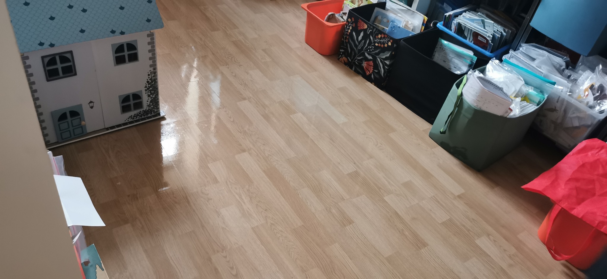 Błyszcząca podłoga po umyciu, z widocznymi pojemnikami z zabawkami i pudełkami z rzeczami, w rogu pokoju stoi domek dla lalek.