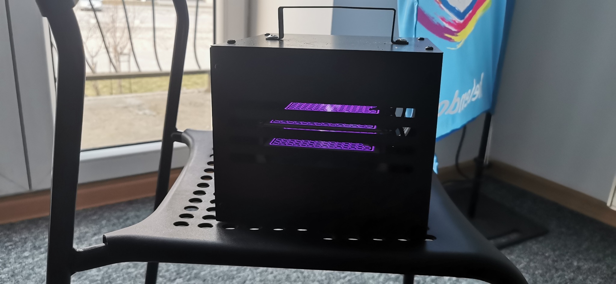 Czarna, metalowa skrzynka z uchwytem na czarnym krześle, z widocznymi fioletowymi światłami przez otwory wentylacyjne.