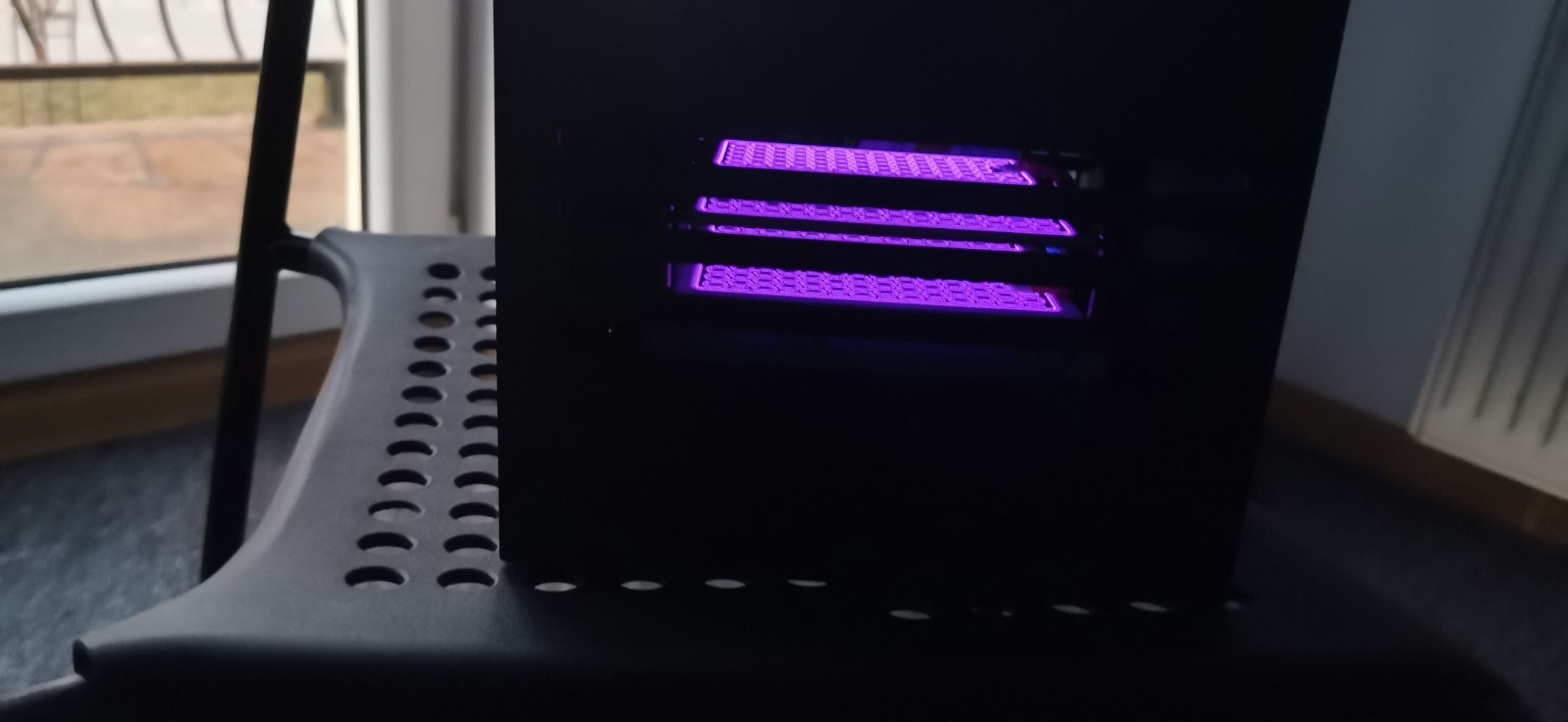 Czarna obudowa urządzenia z trzema poziomami siatki oświetlonej fioletowym światłem, stojąca na czarnym krześle z perforowanym siedziskiem, w tle widoczne okno.
