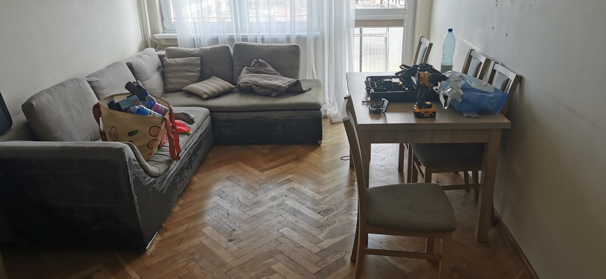 Pokój po remoncie: narożna kanapa z torbą na zakupy, stół z wiertarką i narzędziami, ślady po wierceniu na ścianie, parkiet w jodełkę.