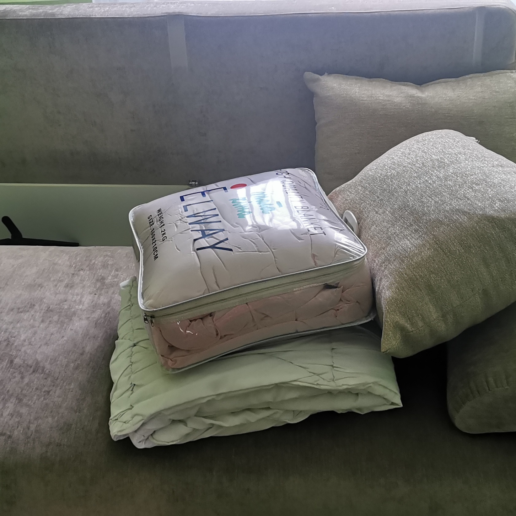 Zapakowany komplet pościeli marki ELWAY, w przezroczystym opakowaniu z suwakiem, leży na złożonej, jasnozielonej kołdrze na ciemnoszarej kanapie obok poduszek.
