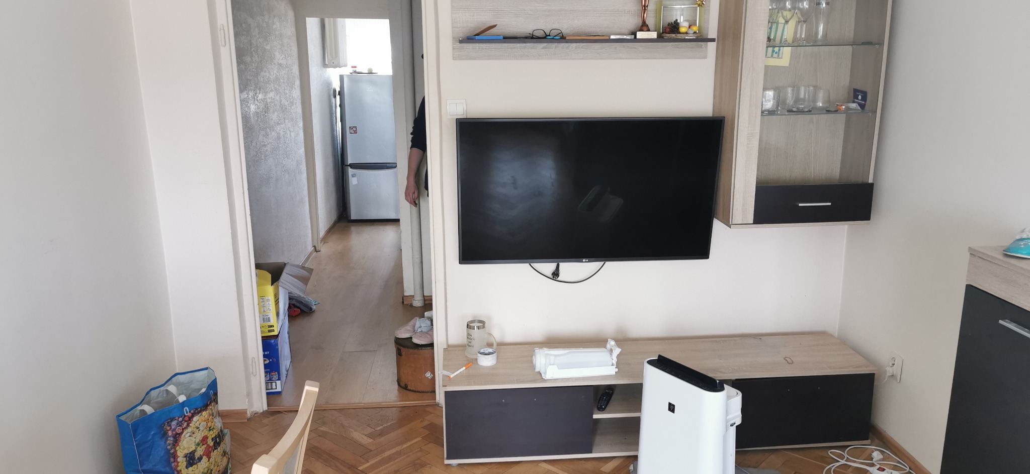 Salon w trakcie porządkowania: widoczny telewizor na ścianie, szafka RTV, oczyszczacz powietrza, kartony i torba na podłodze, oraz otwarte drzwi do kuchni z lodówką.