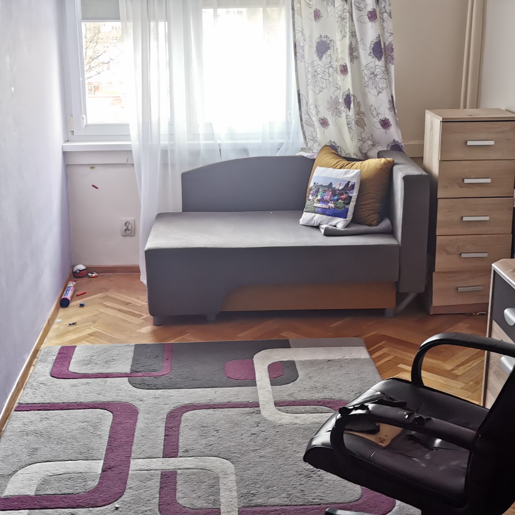 Pokój z szaro-żółtą sofą, drewnianą komodą, dywanem w geometryczne wzory i zabawkami na podłodze.