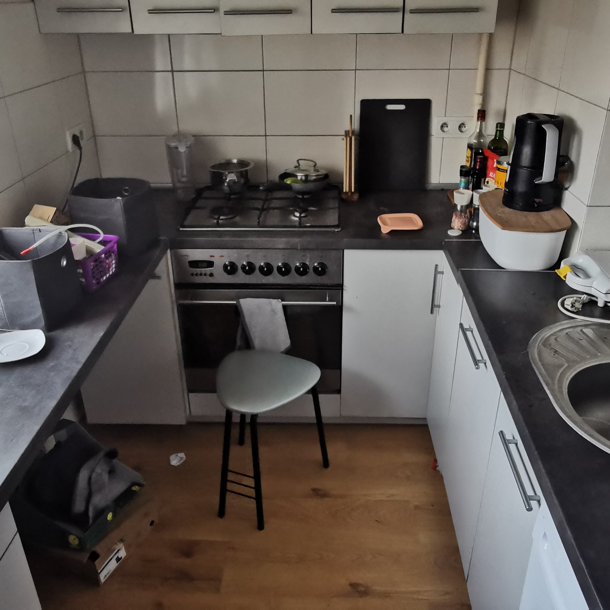 Kuchnia z wyposażeniem, w tym kuchenka gazowa, piekarnik, czajnik elektryczny, zlew i szafki. Na blacie widoczne naczynia i przyprawy. Krzesło stoi przed piekarnikiem.