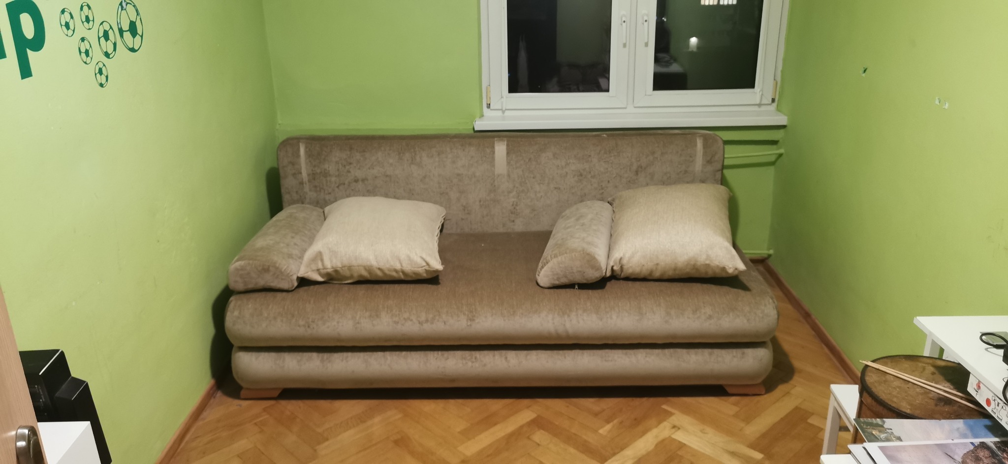 Beżowa sofa z poduszkami w pokoju z zielonymi ścianami i drewnianą podłogą, widoczne naklejki piłkarskie na ścianie.