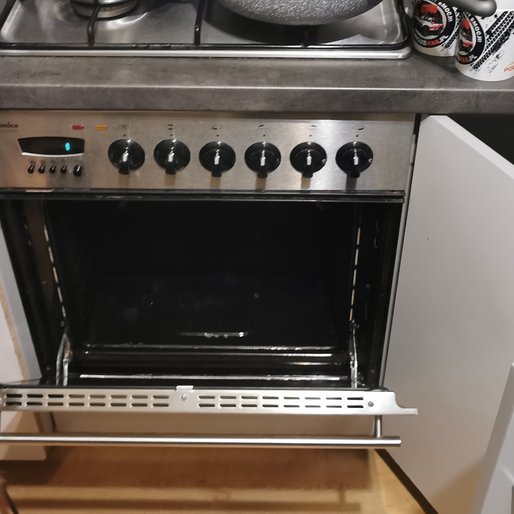 Otwarty piekarnik Amica z widocznymi zabrudzeniami wewnątrz, obok stoją kubki z motywem offroad.