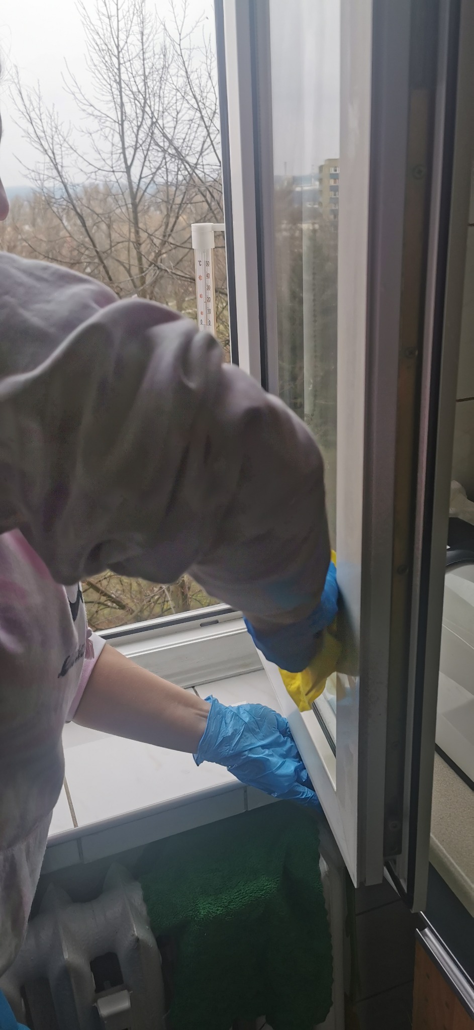 Osoba w niebieskich rękawiczkach czyści ramę okienną żółtą gąbką, widoczny termometr na oknie i fragment grzejnika pod spodem.