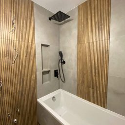 Oliwia Domagalska - Nowoczesna łazienka z drewnianymi panelami ściennymi, szarą płytką, białą wanną i czarną armaturą, widoczne niedokończone podłączenia instalacji.