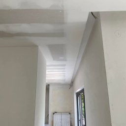 Oliwia Domagalska - Wnętrze w trakcie prac wykończeniowych: sufit z płyt kartonowo-gipsowych z widocznymi łączeniami, ściany przygotowane pod malowanie, widoczne przewody elektryczne i grzejnik zabezpieczony folią.