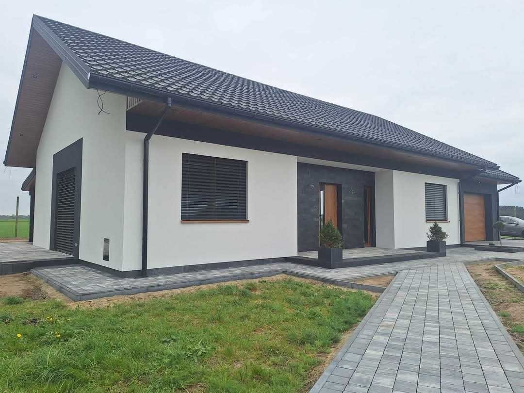 Nowoczesny dom jednorodzinny z białymi ścianami, grafitowym dachem i elementami z ciemnego kamienia, widoczny z zewnątrz, z chodnikiem z kostki brukowej prowadzącym do wejścia.